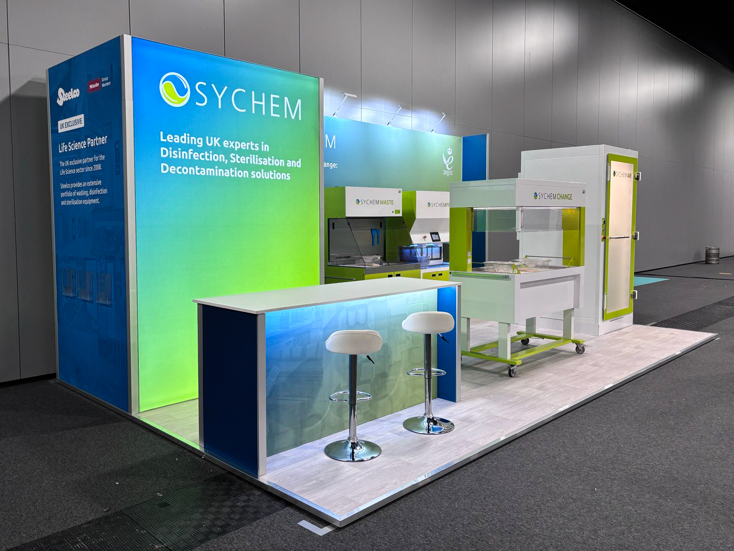 large exhibition stand with blue and green walls at the back. A lit counter with two stools and some chemical equipment are set in front of the rear wall